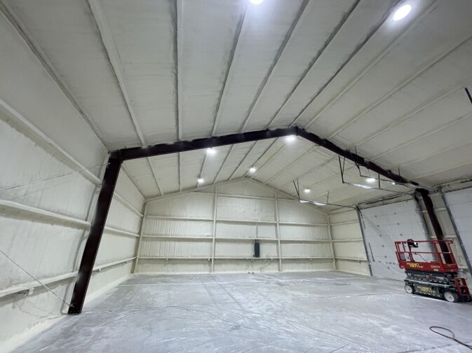 Large warehouse interior with white spray foam insulation on walls and ceiling, exposed steel beams, plastic covering the floor, and a red scissor lift on the right side.