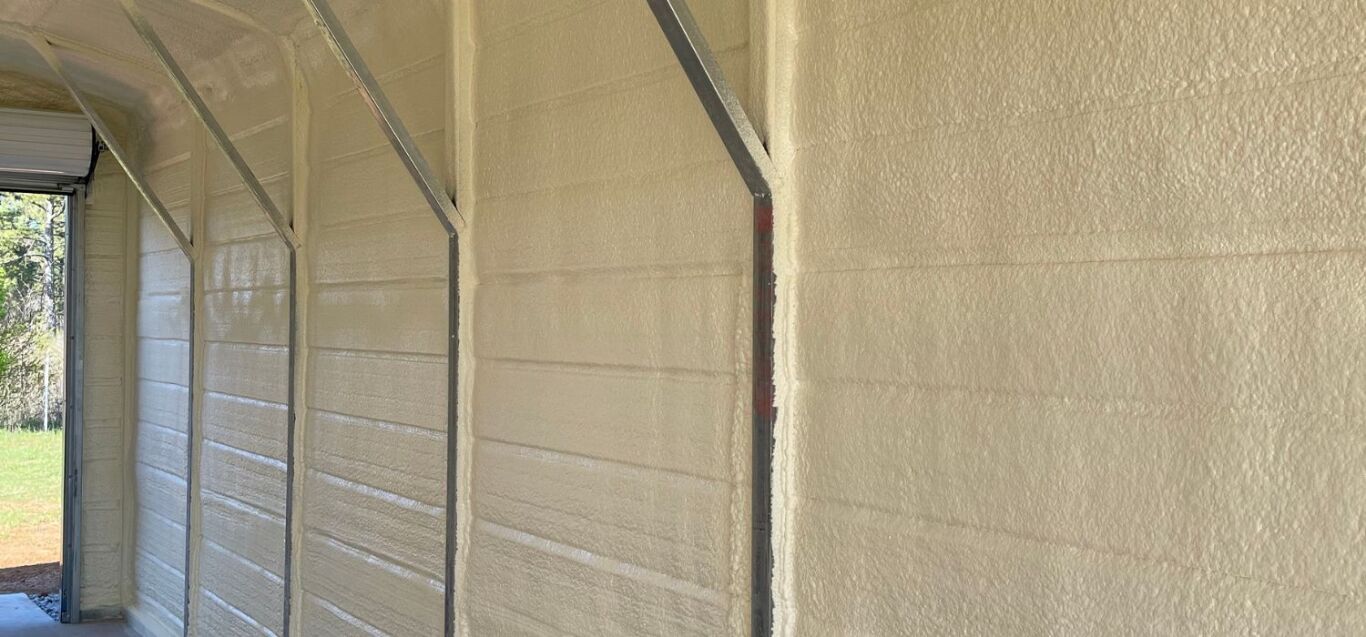 A garage interior wall covered with spray foam insulation, showing metal support beams and an open door letting in natural light.