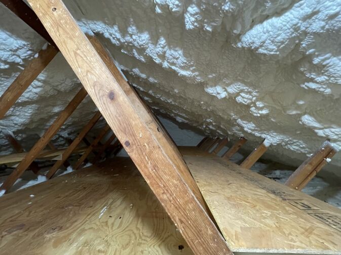 Attic interior with wooden beams and plywood flooring, showing spray foam insulation applied to the underside of the roof.
