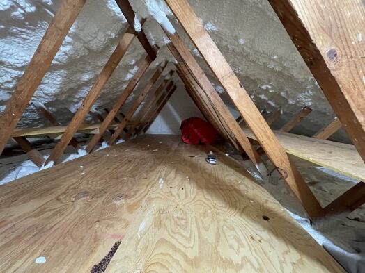 Unfinished attic with wooden beams, spray foam insulation on the ceiling, plywood flooring, a red bag, and a few small items on the floor.