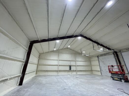 Large warehouse interior with white spray foam insulation on walls and ceiling, exposed steel beams, plastic covering the floor, and a red scissor lift on the right side.
