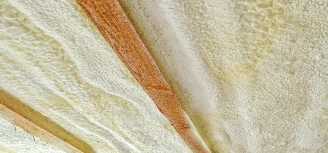 Close-up view of a ceiling with exposed wooden beams and spray foam insulation applied between them.