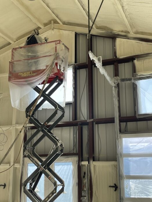 A scissor lift holds equipment near the ceiling inside a building under construction, with insulation and protective plastic covering walls and windows.