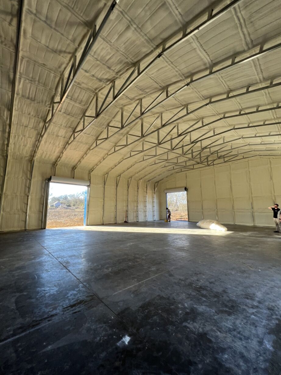 Wide-angle view of the interior of an empty warehouse with high ceilings, exposed insulation, open doors, and two people standing near the far wall.