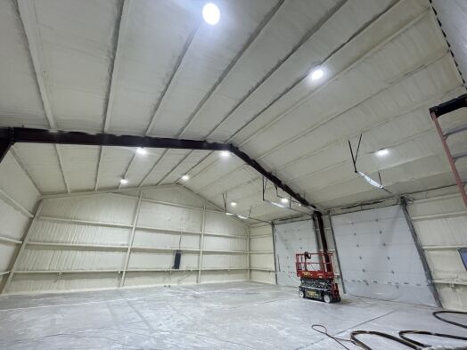Large industrial interior with white spray foam insulation on walls and ceiling, overhead lights, a red scissor lift, and a large garage door.