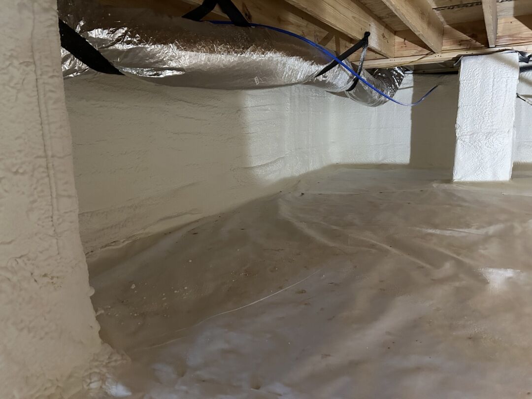 A crawl space with spray foam insulation on the walls, plastic vapor barrier on the ground, and visible ductwork and wooden beams above.