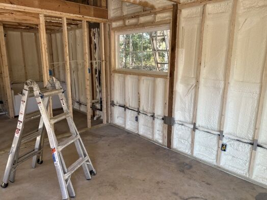 A room under construction with exposed wooden framing, foam insulation in the walls, a ladder, and a window letting in natural light.