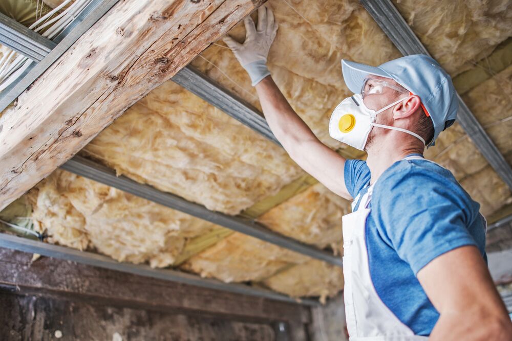 A worker wearing protective gear performs insulation removal and installs new insulation material on the ceiling of a building.