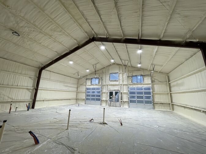 Interior of a large building under construction with spray foam insulation on walls and ceiling, exposed steel beams, and plastic covering the floor. Multiple garage doors and windows are visible.