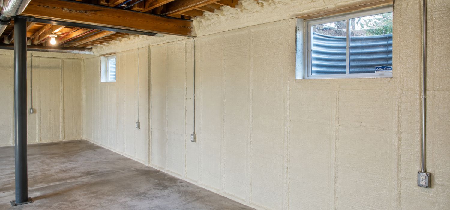Basement with spray foam insulation on the walls, exposed ceiling beams, concrete floor, metal support post, electrical outlets, and two small windows with window wells—showcasing quality basement insulation in Huntsville.