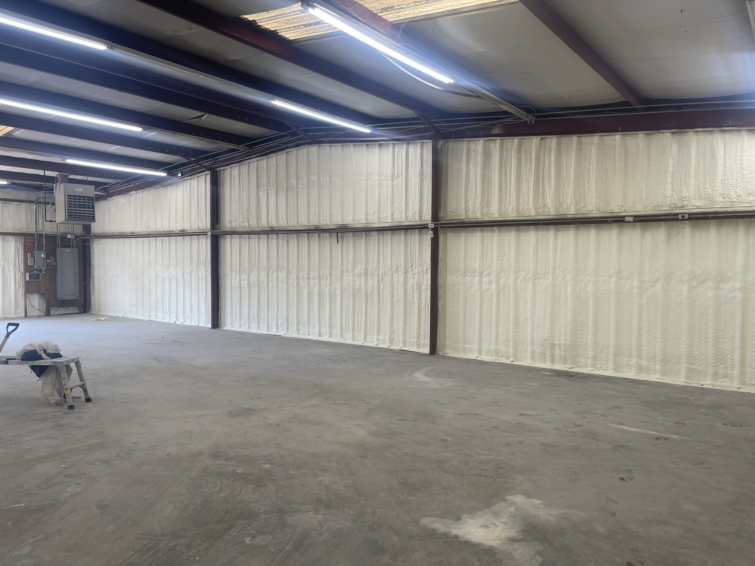 Interior of an industrial building with spray foam insulation applied to the walls, exposed steel beams, fluorescent lights, and a few tools on the concrete floor.