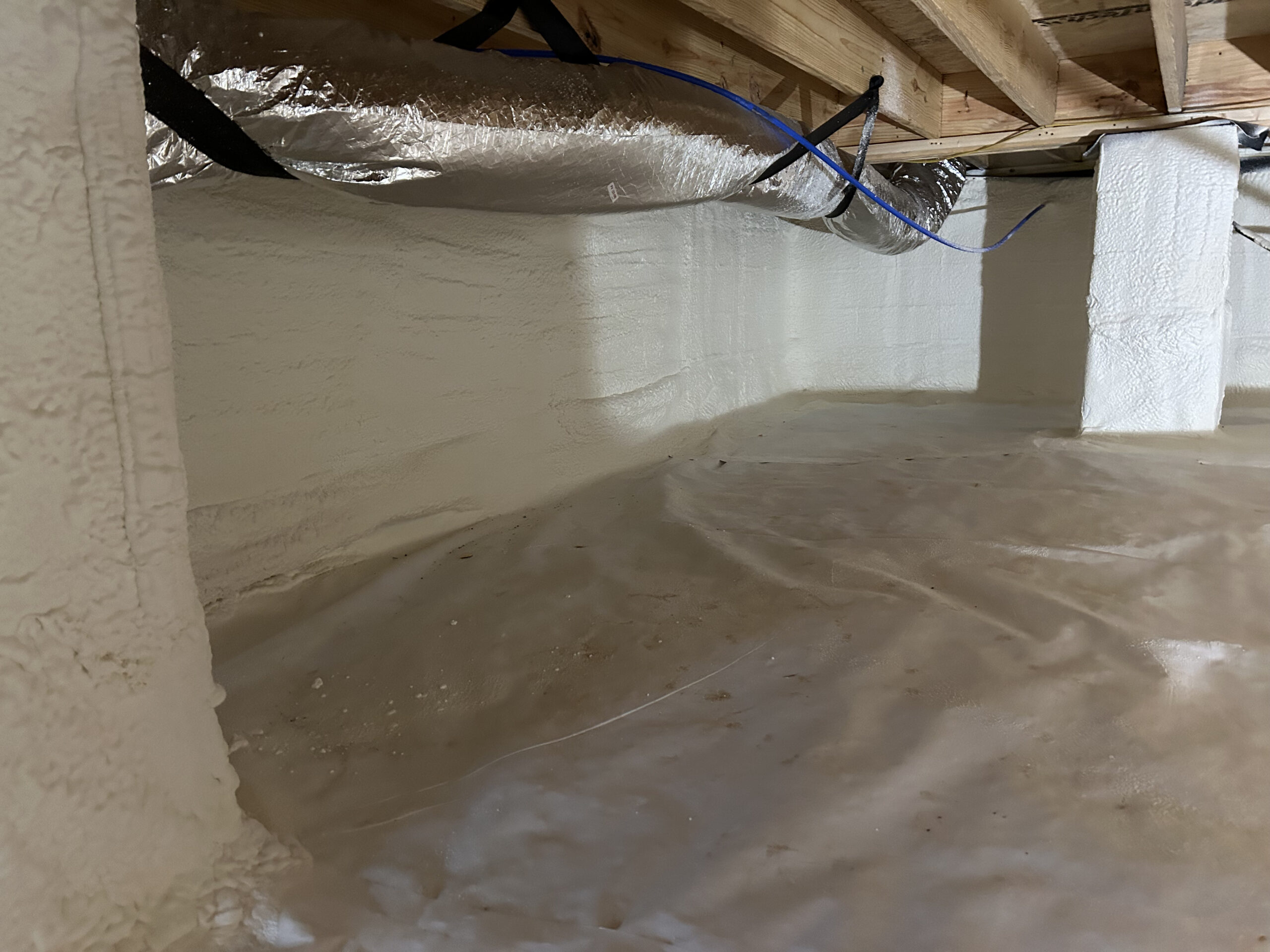 A crawl space with spray foam insulation on the walls, plastic vapor barrier on the ground, and visible ductwork and wooden beams above.
