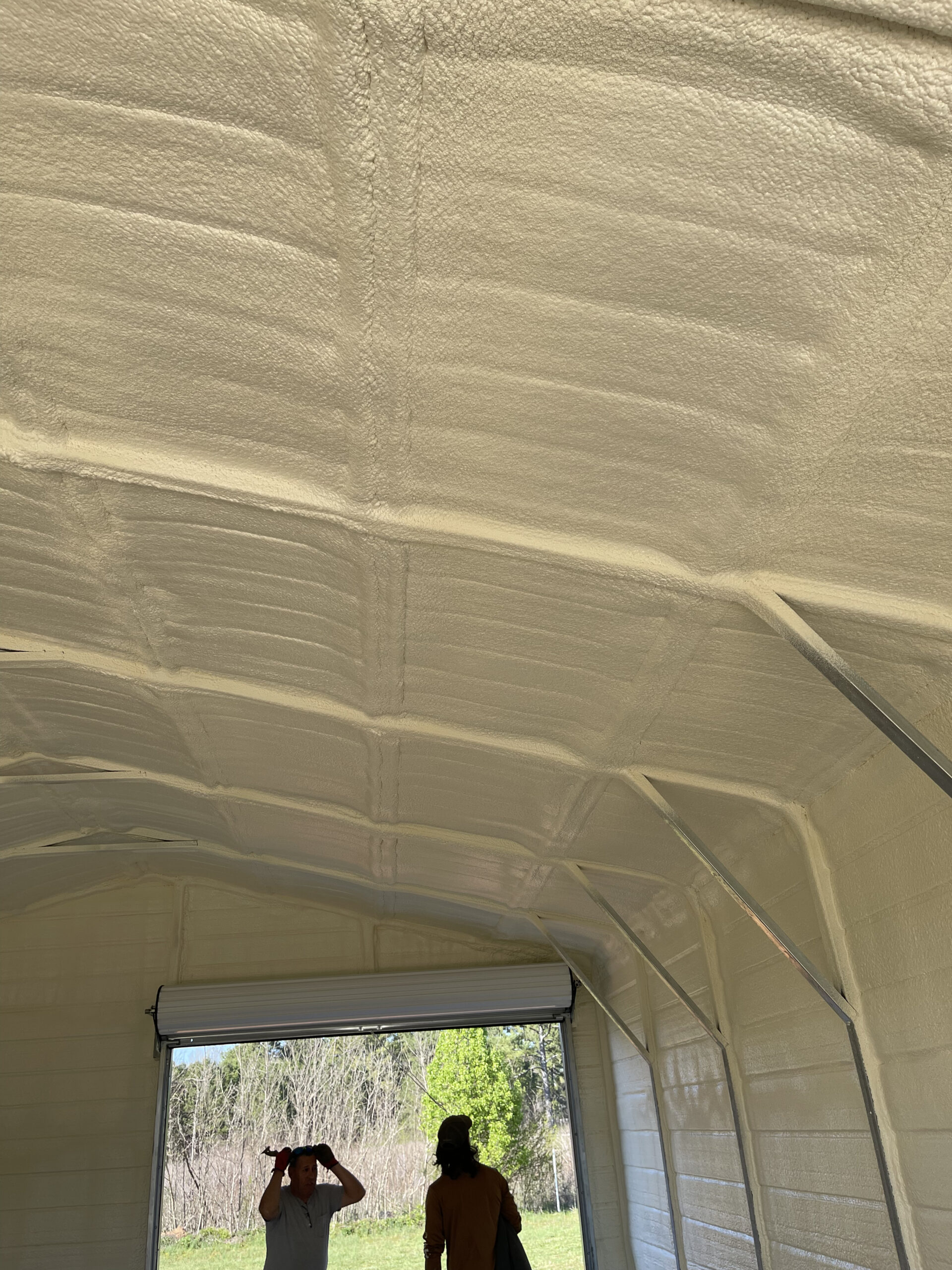 Two people stand inside a building with white spray-foam insulated walls and ceiling, looking out an open garage door toward a green outdoor area.