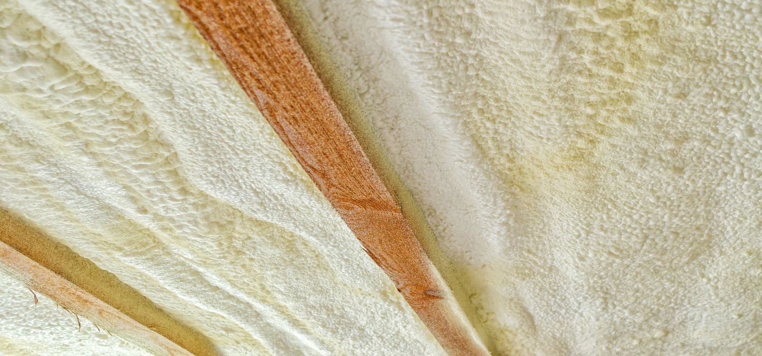 Close-up view of a ceiling with exposed wooden beams and spray foam insulation applied between them.