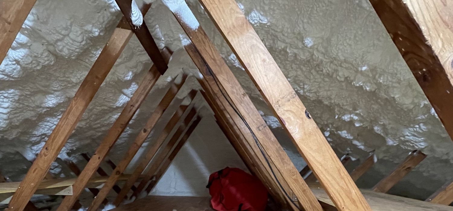 Spray foam insulation applied along the roofline of an older home's attic, sealing gaps between wooden rafters to improve energy efficiency.