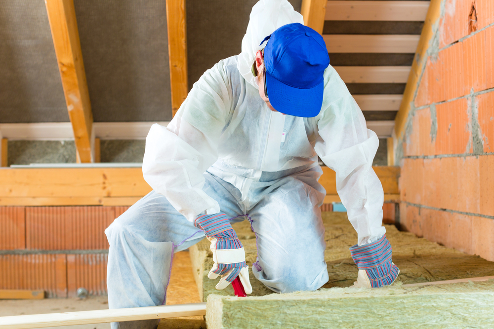 Person in protective clothing and gloves performs insulation removal and installs new material in an attic with exposed wooden beams and brick walls.