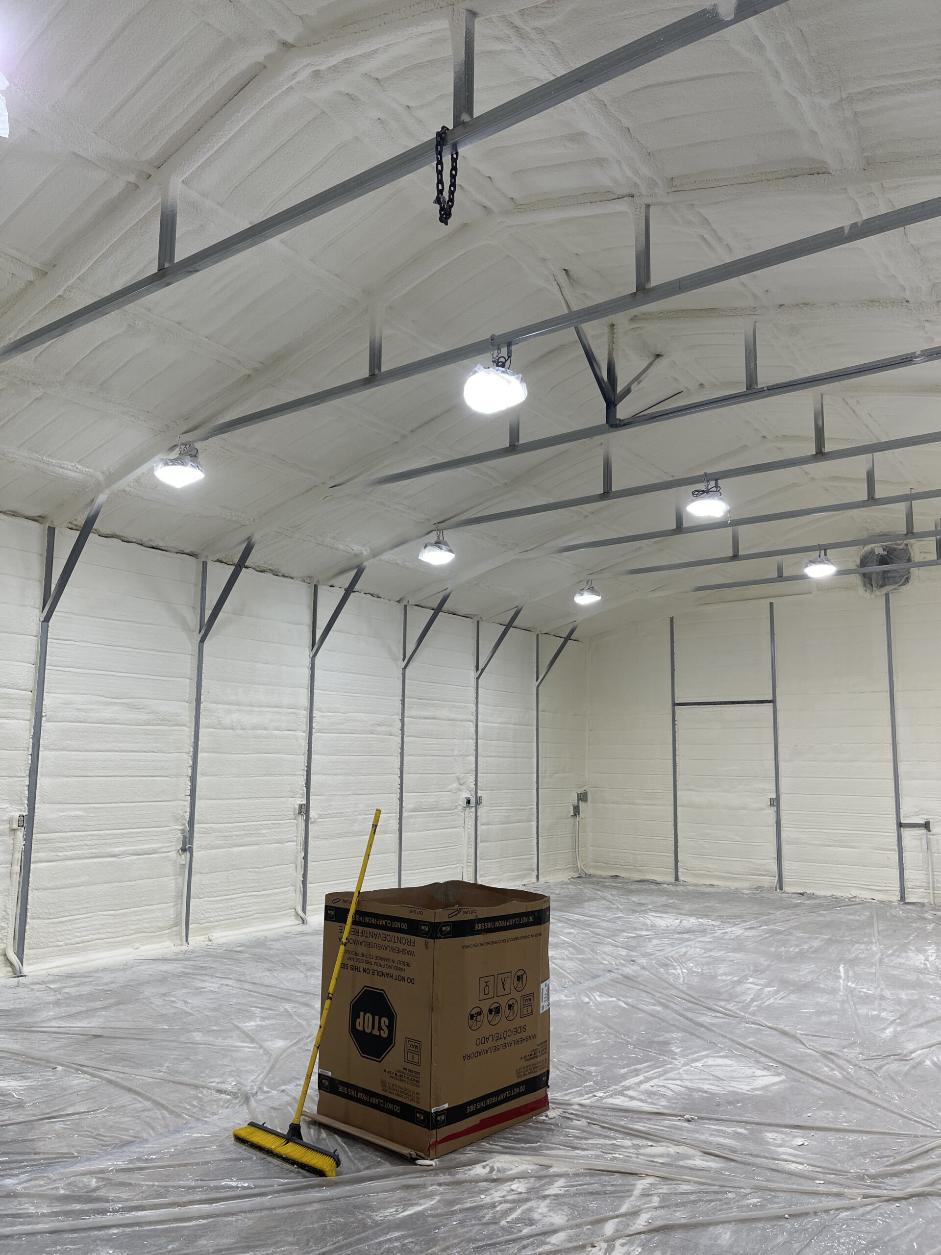 A large, well-lit industrial space with spray foam insulation on the walls and ceiling, a broom, and a cardboard box on a plastic-covered floor.