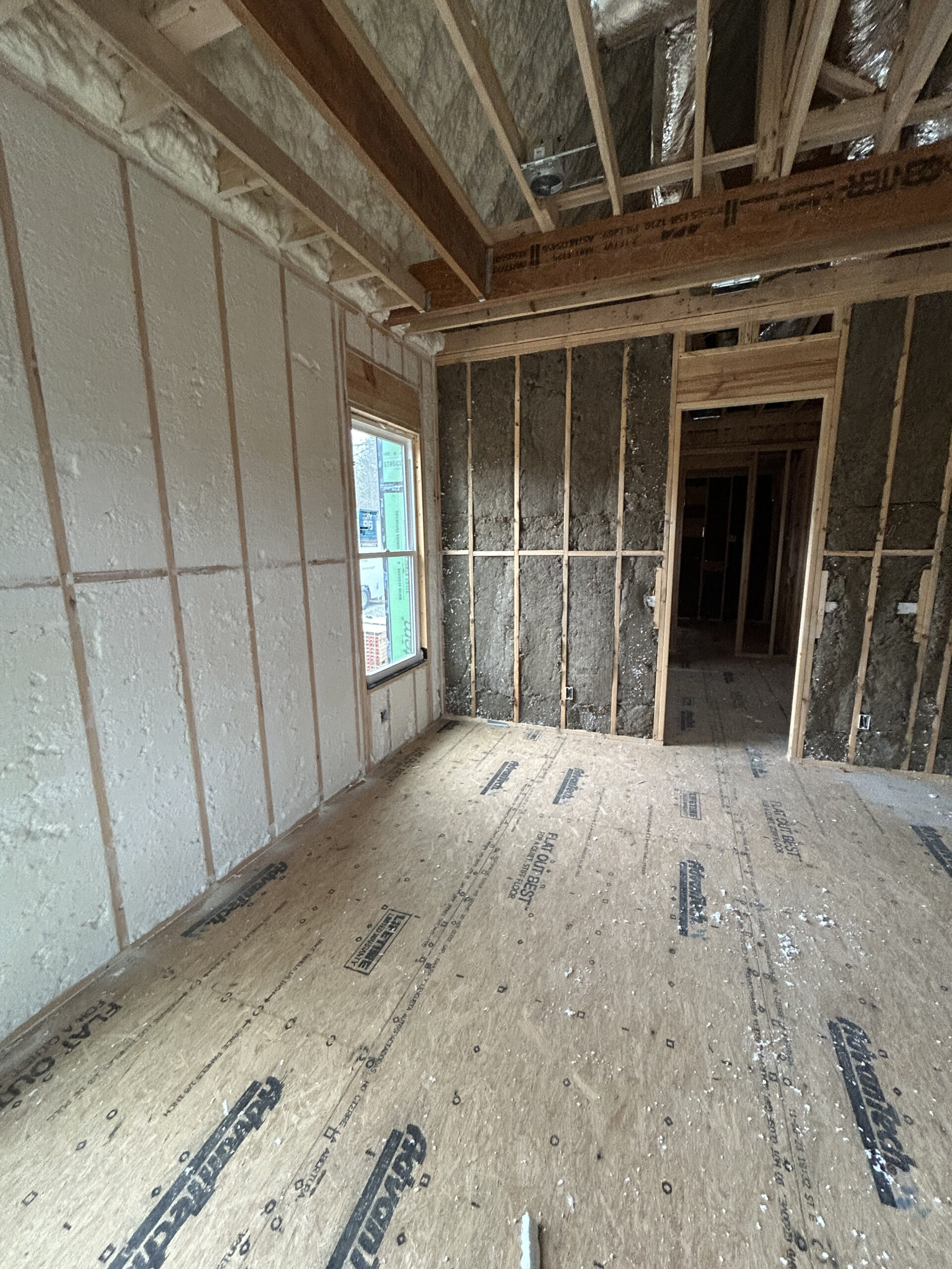 Room under construction with exposed wood framing, spray foam insulation on some walls, and an unfinished floor. Window and doorway openings visible.