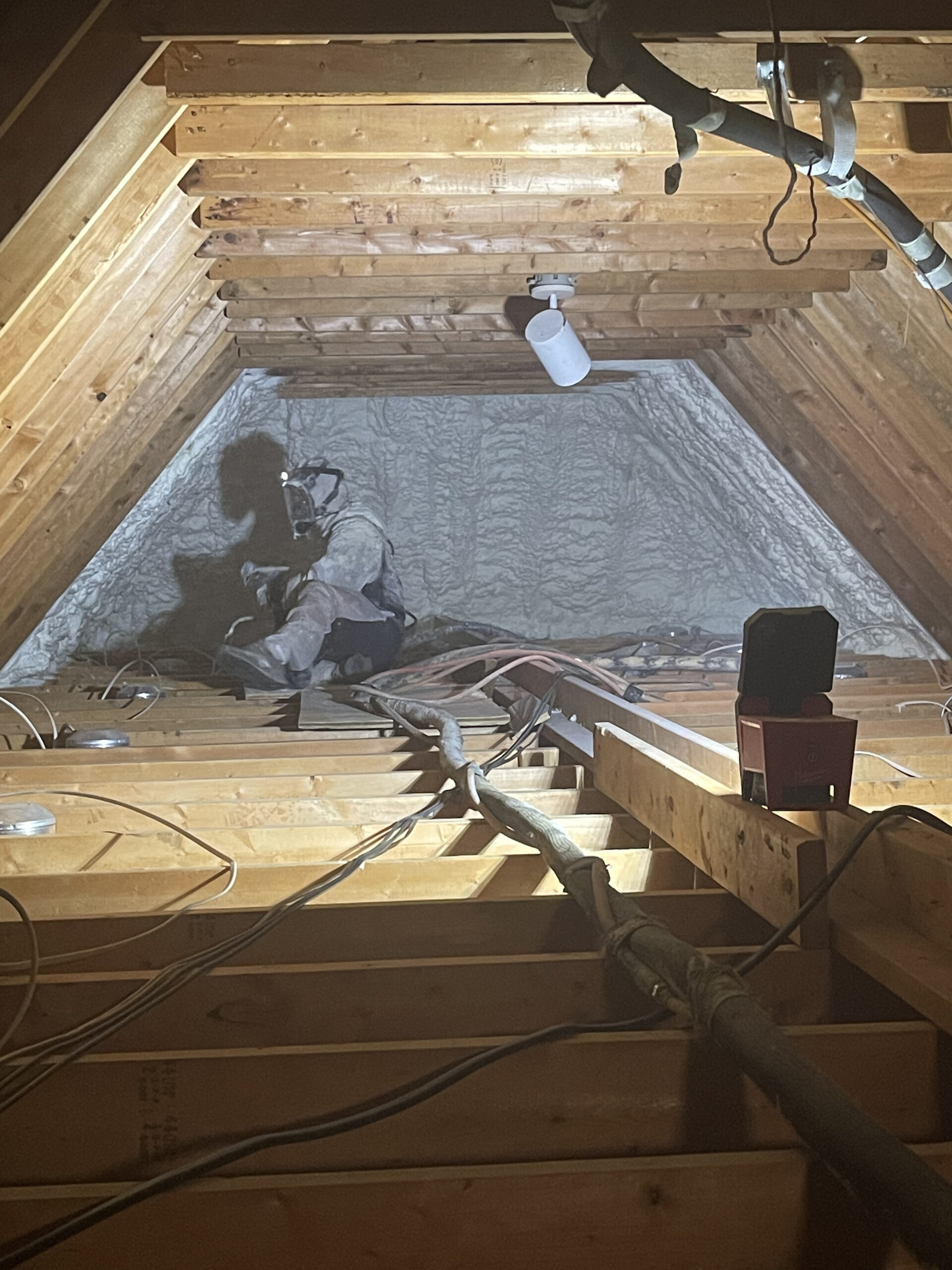 Worker in protective gear sprays foam insulation on the far wall of a wooden attic with exposed beams and wiring.
