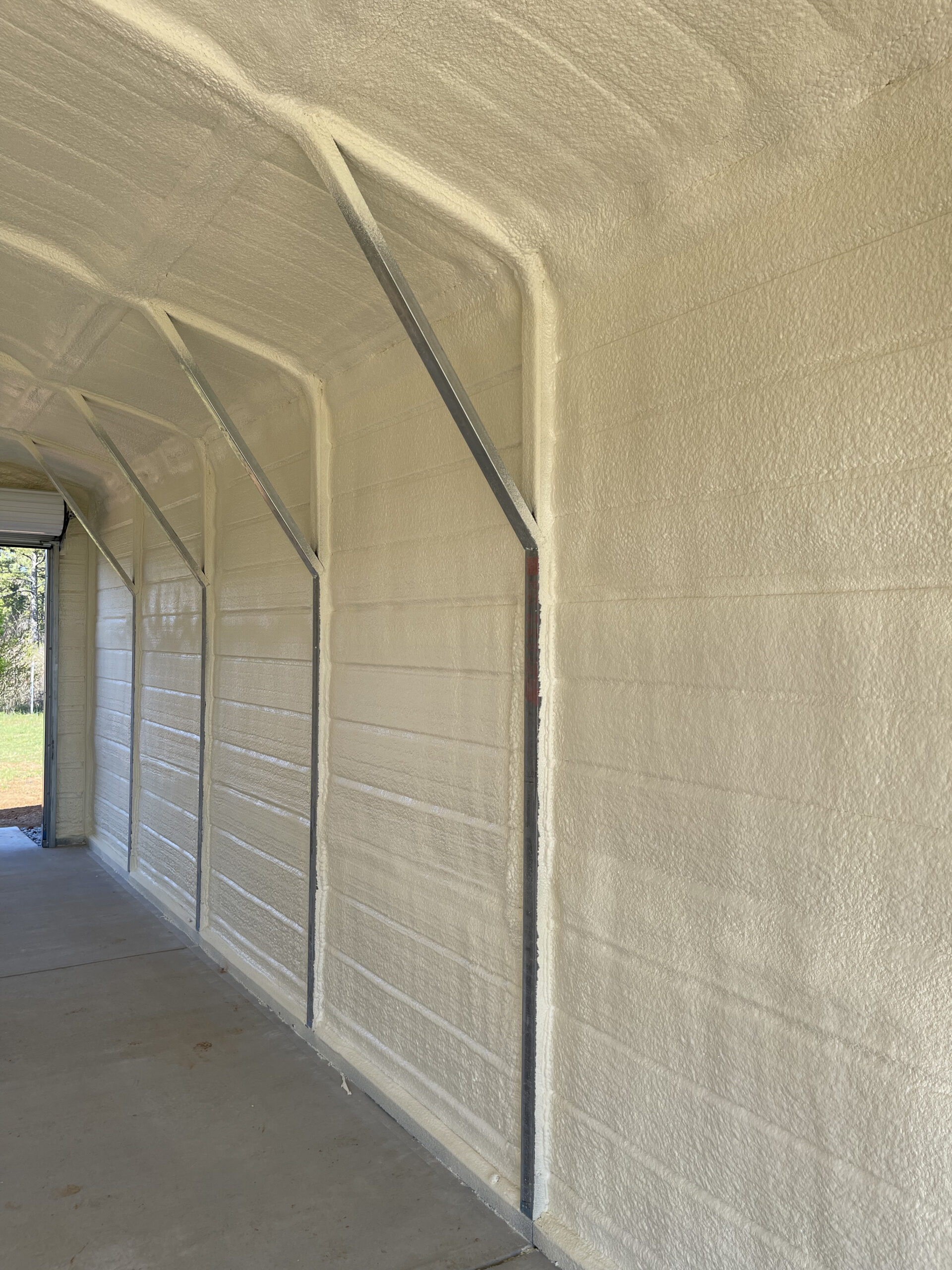 Interior of a metal building with spray foam insulation applied to the walls and ceiling, showing exposed steel supports and an open entrance at one end.