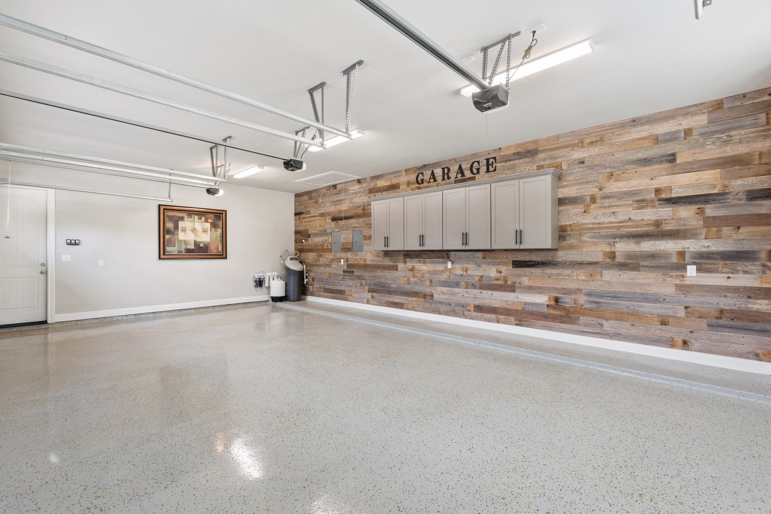 Spacious, clean garage with polished speckled floor, wood accent wall, cabinets, and a framed artwork. Ceiling-mounted garage door mechanisms are visible.