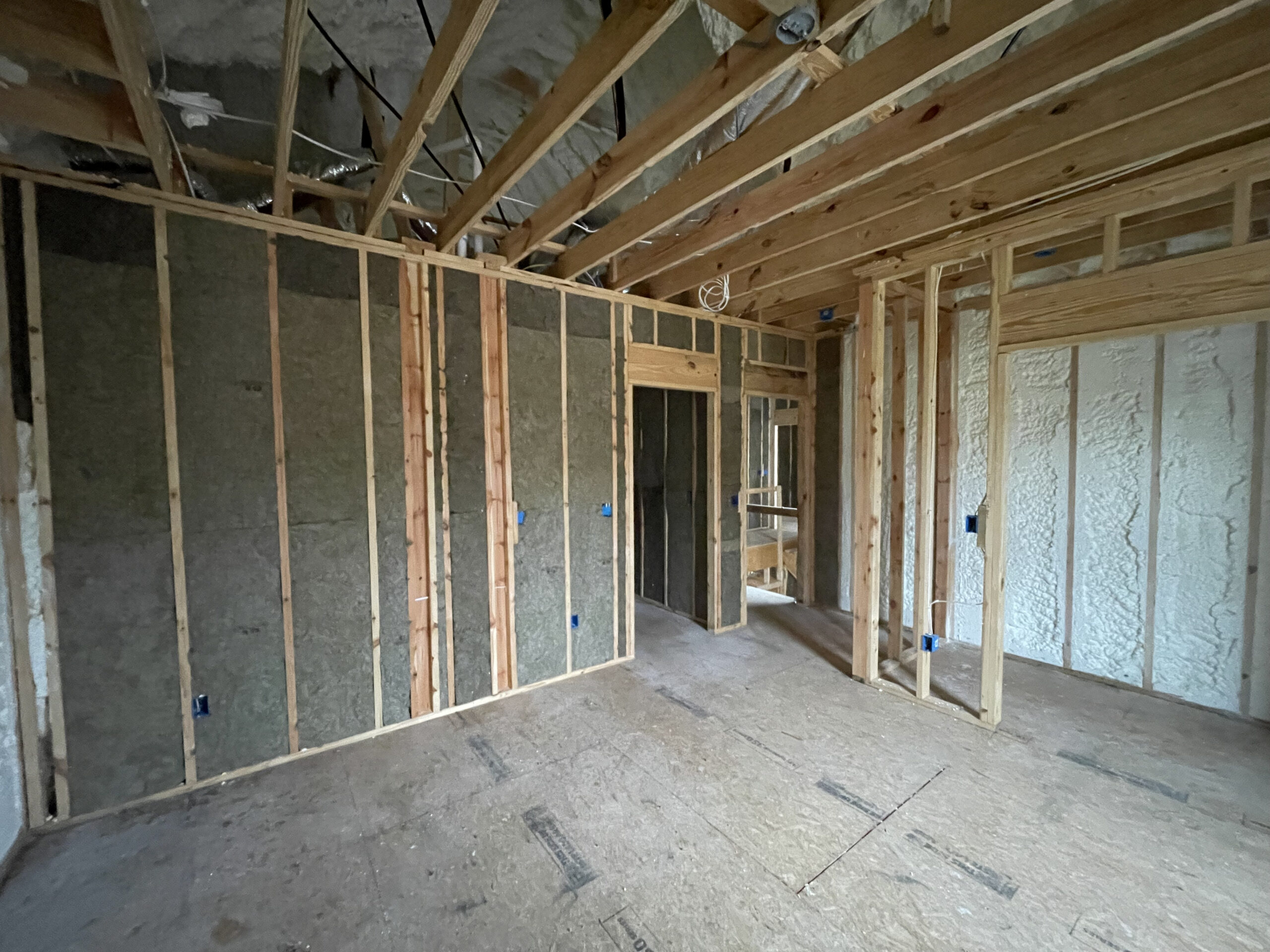 A room under construction with exposed wooden framing, insulation in the walls, electrical boxes installed, and an unfinished floor and ceiling.